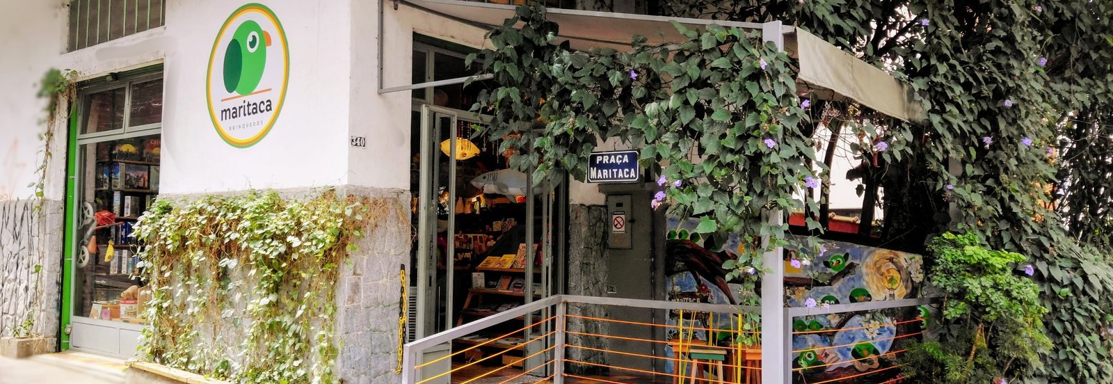 Fachada da Maritaca Brinquedos na Rua Paracuê: parede branca com o logotipo, vitrine iluminada, trepadeiras e a varanda Praça Maritaca